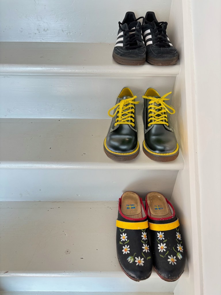 Three pairs of shoes placed on white wooden steps. A pair of German Adidas trainers, English Doc Martens and Swedish clogs.