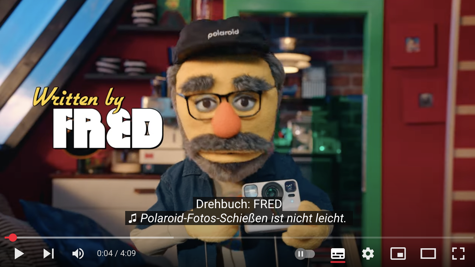 Still of puppet in a studio environment holding up a Polaroid camera. The superimposed title says "Written by Fred". Subtitles read 'Drehbuch: FRED' - Directed by FRED. A song is audible with the lyrics 'Polaroid pics can be tricky to take.' German subtitles read: "Polaroid-Fotos-Schießen ist nicht leicht." 